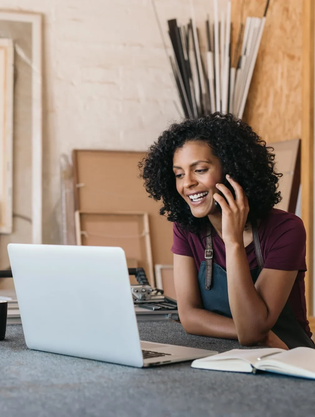 Happy woman talking on phone while working on laptop in creative workspace.